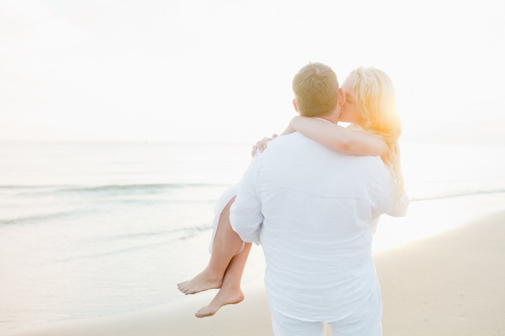 Photo groom carries bride into sunset alongside the ocean